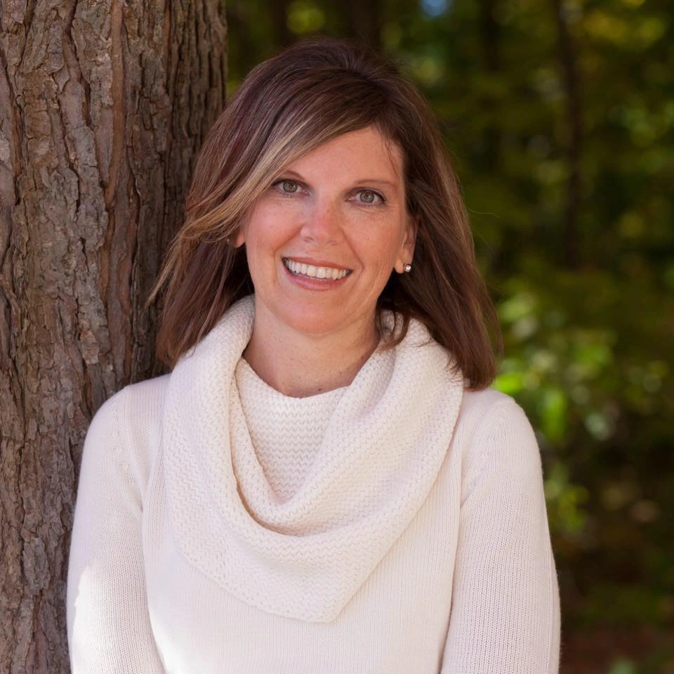 Elizabeth standing by a tree in an off-white sweater with cowl neck. Elizabeth is smiling